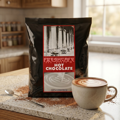 CAFFEIN8 Hot Chocolate Mix Powder 2 lb bag on a kitchen counter beside a ceramic mug of hot chocolate with latte art, cocoa powder, and a spoon, styled for office breakroom use